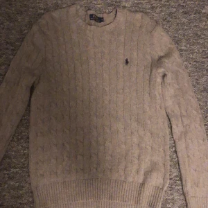 Beige stickad tröja från Ralph Lauren - Säljer en stilren beige stickad tröja från Ralph Lauren med kabelstickat mönster. Tröjan har långa ärmar och en liten broderad logotyp på bröstet. Perfekt för kyliga dagar!