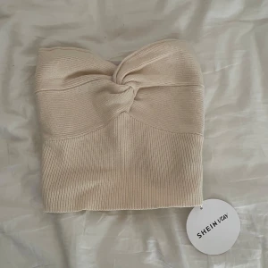 Beige ribbad tubtopp från SHEIN - Säljer en stilren beige tubtopp från SHEIN. Toppen är ribbad och har en knutdetalj framtill som ger en unik touch. Perfekt för en trendig look.