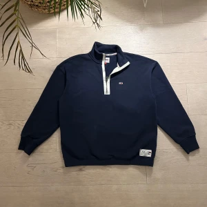 Mörkblå tröja från Tommy Jeans - Säljer en snygg mörkblå tröja från Tommy Jeans med dragkedja och liten logga på bröstet. Tröjan har långa ärmar och en stilren design, perfekt för en casual look. Passar bra till jeans eller chinos.