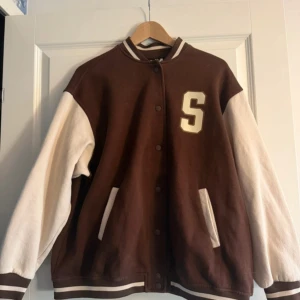 Brun och vit bomberjacka med bokstavsdetalj - Snygg brun och vit bomberjacka med ett stort 'S' på framsidan och 'Seattle' på baksidan. Jackan har långa ärmar och stängs med knappar. Perfekt för en cool och avslappnad stil.