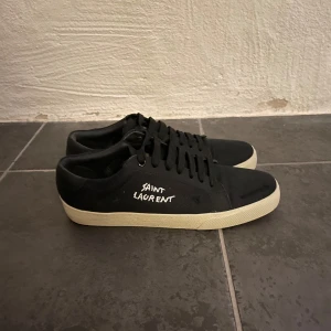 Svarta sneakers från Saint Laurent - Snygga svarta sneakers från Saint Laurent med vit sula och märkets logga i vitt på sidan. Skorna har klassisk snörning och en stilren design som passar perfekt för en trendig look. Anvönda några gånger men säljer eftersom de inte passar mig 