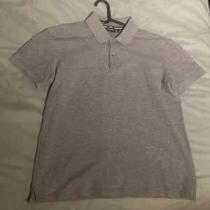 Grå pikétröja från J.Lindeberg - Snygg grå pikétröja från J.Lindeberg med klassisk krage och knappar framtill. Perfekt för en stilren look. Tröjan har korta ärmar och är tillverkad i ett bekvämt material.