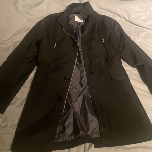 Svart kappa från J.Lindeberg - Elegant svart kappa från J.Lindeberg med knappar och dragkedja framtill. Jackan har långa ärmar och en stilren design med ett bälte baktill för justerbar passform. Perfekt för en stilfull look.