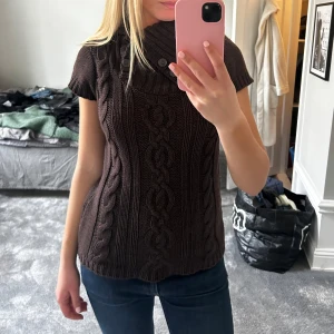 Brun stickad tröja med knappar - Säljer en brun stickad tröja med korta ärmar och en hög krage dekorerad med knappar. Tröjan har ett snyggt flätmönster som ger en stilren look. Perfekt för kyligare dagar!