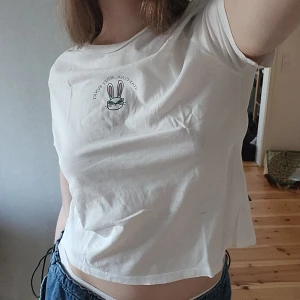 Vit t-shirt med tryck - Säljer en vit t-shirt med ett tryck av en kanin och texten 'Stay Cool, Honey Bunny'. T-shirten är lite croppad och har och korta ärmar. Perfekt för en avslappnad stil.