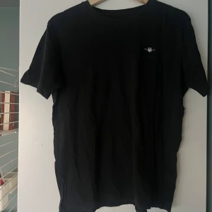 Svart t-shirt från GANT - Säljer en klassisk svart t-shirt från GANT. Den har en liten logga på bröstet och är tillverkad i ett mjukt material. Perfekt för en stilren look.
