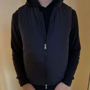 Svart dunväst från Oscar Jacobson - Snygg svart dunväst från Oscar Jacobson med dragkedja framtill. Västen är vindtät, vattenavvisande och andas bra, vilket gör den perfekt för olika väderförhållanden. Den har en stilren design och passar bra över en hoodie eller tröja.Använd ett fåtal gånger 9/10 skick. NYPRIS 1999kr