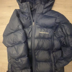 Mörkblå pufferjacka från Peak Performance - Säljer en snygg mörkblå pufferjacka från Peak Performance. Jackan har en dragkedja framtill och en praktisk huva. Perfekt för kyliga dagar och håller dig varm och bekväm.
