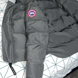 Grå dunjacka från Canada Goose - Säljer en stilren grå dunjacka från Canada Goose med deras ikoniska märke på ärmen. Jackan har en dragkedja framtill och är perfekt för kalla dagar. Den är långärmad och har en praktisk huva för extra värme.