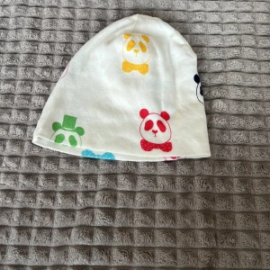 Minirodini mössa - Söt vit mössa med färgglada pandor i gult, grönt, rött och blått. Perfekt för att liva upp vilken outfit som helst!