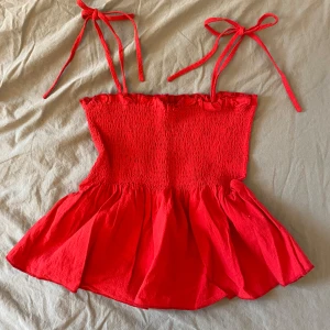 Röd ärmlös topp med knytband - Snygg röd topp med smockdetaljer och volangkant. Den har knytband vid axlarna och en luftig passform nedtill. Perfekt för varma dagar!
