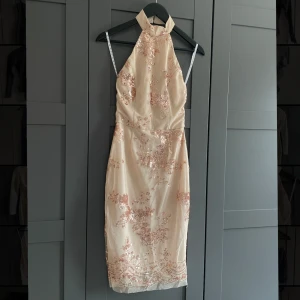 Paljettklänning med öppen rygg - Champagnefärgad klänning i midi-modell med rosafärgade glittrande paljetter i blommigt mönster. Klänningen har en hög hals med två knappar baktill. Öppen rygg och dragkedja baktill.