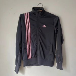  träningsjacka från Adidas - Snygg träningsjacka från Adidas med rosa detaljer och de klassiska tre ränderna på sidan. Jackan har dragkedja och långa ärmar, perfekt för träning eller en sportig look. Adidas-loggan är broderad i rosa på bröstet. Har några fläckar som jag ej provat att få bort (se 2 sista bilderna)⭐️