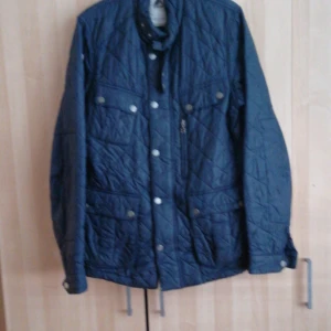 Blå quiltad jacka från 8848 Altitude - Snygg blå quiltad jacka från 8848 Altitude med knappar och dragkedja. Jackan har långa ärmar och flera fickor med knappstängning. Perfekt för kyligare dagar.