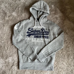 Superdry hoodie - Säljer en grå hoodie från Superdry med tryck i blått och vitt. Tröjan är i toppskick och knappt använd. Inga defekter.