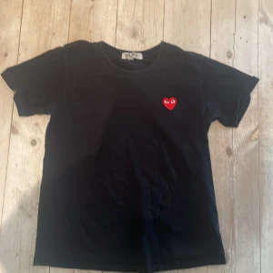 Comme des Garçons t-shirt  - Säljer en stilren svart t-shirt från Comme des Garçons med ett ikoniskt rött hjärta på bröstet. Perfekt för en minimalistisk look. Kortärmad och tillverkad i mjukt material för extra komfort.
