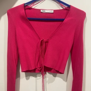 Råsa ribbad topp från Zara - Säljer en snygg rosa ribbad topp från Zara med v-ringning och knytning framtill. Toppen är croppad och har långa ärmar, perfekt för en trendig look. Passar bra till både jeans och kjol. Helt oanvänd!
