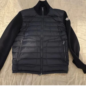 Svart dunjacka från Moncler - Snygg svart dunjacka från Moncler med dragkedja och långa ärmar. Jackan har en quiltad design och Moncler-logga på ärmen. Perfekt för kyligare dagar eller till börsen våren