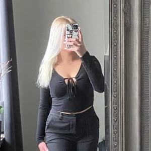 Svart ribbad topp med knytning - Snygg svart ribbad topp med lång ärm och knytning framtill. Perfekt för en stilren look. Toppen har en v-ringad design som ger en elegant touch.