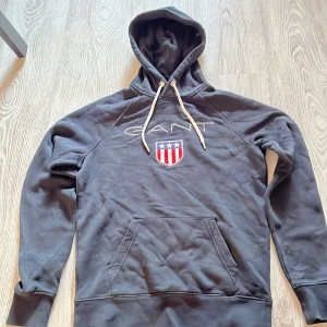 Svart hoodie från GANT - Säljer en svart hoodie från GANT med en stor ficka fram och justerbar huva. Den har ett broderat emblem på bröstet och vita snören. Perfekt för en avslappnad stil.Storlek XS.