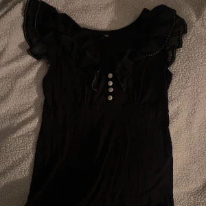 Svart blus med volangdetaljer - Snygg svart topp med volangdetaljer vid axlarna och knappar framtill. Aldrig använd 