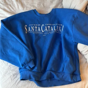 Blå sweatshirt från Gina Tricot - Säljer en blå sweatshirt från Gina Tricot med texten 'Santa Catalina Fitness Institution' tryckt på framsidan. Tröjan har långa ärmar och ribbade muddar vid ärmslut och nederkant. Perfekt för en avslappnad stil till sommaren