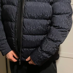 Svart mönstrad dunjacka från Moncler - Säljer en snygg svart dunjacka från Moncler med ett diskret mönster. Jackan har en dragkedja framtill och Moncler-logga på ärmen. Perfekt för kyliga dagar med sin vadderade design.