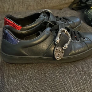 Svarta sneakers från Gucci - Snygga svarta sneakers från Gucci i skinn med unika detaljer. Skorna har en dekorativ spänne på sidan och färgade hälpartier i rött och blått. Perfekta för dig som vill ha något extra i garderoben. Knappt använda. Nypris 6000kr.