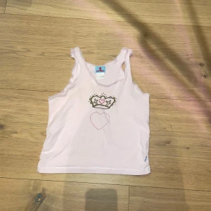 Vit linnetopp från CCC Girls - Säljer en söt vit linnetopp från CCC Girls med ett tryck av en krona och ett hjärta på framsidan. Perfekt för en avslappnad stil. Toppen är ärmlös och har en enkel design.