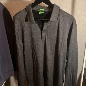 Grå pikétröja från BOSS - Säljer en stilren grå pikétröja från BOSS med modern fit. Tröjan har långa ärmar och en klassisk krage med knappar framtill. Perfekt för en avslappnad men ändå elegant look.