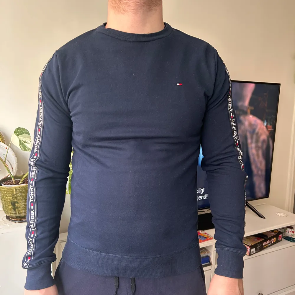 Säljer en stilren mörkblå tröja från Tommy Hilfiger med märkets logga på bröstet och text längs ärmarna. Tröjan har långa ärmar och en rund halsringning. Perfekt för en casual look. Modellen är 185 cm och väger 100 kg. . Neuletakit & Villapaidat.
