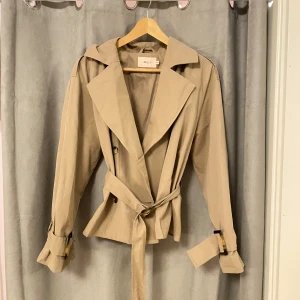 Beige trenchcoat från Nelly - Snygg beige trenchcoat från Nelly med klassisk design. Jackan har ett bälte i midjan och justerbara spännen vid ärmsluten. Perfekt för vår och sommar, säljer då den inte kommer till användning längre tyvärr, väldigt bra skick då den endast är använd några få gånger. ❤️
