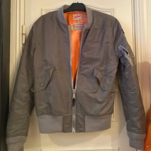 Grön bomberjacka från Schott - Säljer en stilren grön bomberjacka från Schott med orange foder. Jackan har dragkedja framtill och flera praktiska fickor, inklusive en på ärmen. Märkesdetaljer finns på bröstet och ärmen. Perfekt för en cool och avslappnad stil.