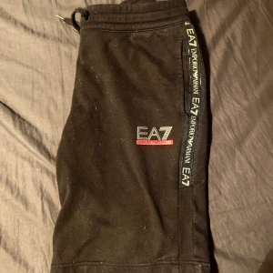 Svarta mjukisshorts från EA7 Emporio Armani - Snygga svarta mjukisshorts från EA7 Emporio Armani med logotypdetaljer längs sidan. Perfekta för en avslappnad stil. De har en elastisk midja med snörning för bästa passform.