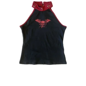 Svart och röd halterneck topp med dödskallemotiv - Snygg svart halterneck topp med röd kant och ett coolt dödskallemotiv på framsidan. Toppen har en hög krage med metallringar och knäppning bak. Perfekt för en edgy look!
