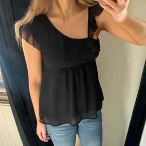 Svart oneshoulder topp med volang - Snygg svart oneshoulder topp med volangdetaljer. Perfekt för en stilren look. Toppen har en luftig passform och är tillverkad i ett lätt material.