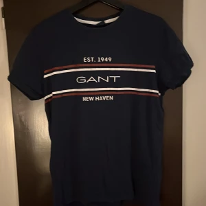 Mörkblå t-shirt från GANT - Säljer en snygg mörkblå t-shirt från GANT med tryck 'EST. 1949 NEW HAVEN' i vitt och rött. T-shirten har en klassisk rund hals och korta ärmar. Perfekt för en avslappnad stil.