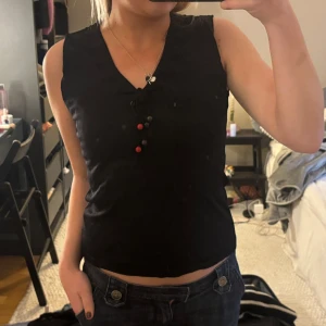 Svart ärmlös topp med knyt och pärlor - Snygg svart ärmlös topp med knytning framtill och dekorativa pärlor i rött och brunt. 