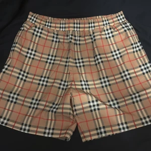 Rutig shorts från Burberry - Snygga rutiga shorts från Burberry i klassiskt beige, svart och rött mönster. De har en elastisk midja för extra komfort och är perfekta för en stilren look. Passar bra till både avslappnade och mer uppklädda tillfällen.