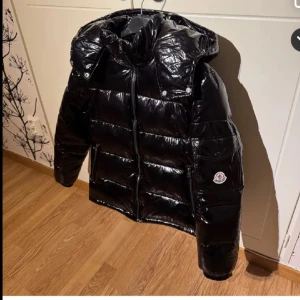 Svart Moncler Maya jacka - Säljer en snygg svart pufferjacka från Moncler. Jackan har en glansig yta med dragkedja och huva. Perfekt för kyliga dagar med sin quiltade design och långa ärmar. En stilren och funktionell jacka för vintern. Passar XS S och M fast den kan vara lite stor på XS. Pris kan diskuteras