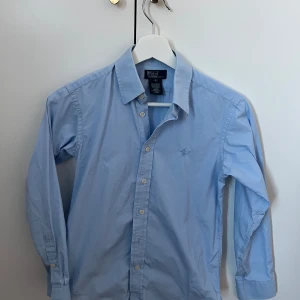 Blå skjorta från Polo Ralph Lauren - Säljer en klassisk ljusblå skjorta från Polo Ralph Lauren. Skjortan har långa ärmar och knappar framtill. Den är broderad med den ikoniska logotypen på bröstet. En knapp är skadad på ärmen och en liten fläck som knappast syns över den broderande loggan.