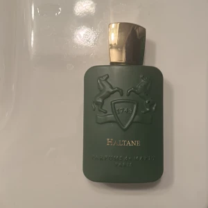 Haltane Parfym från Parfums de Marly - Elegant parfymflaska i mörkgrön med guldlock från Parfums de Marly. Flaskan har en stilren design med upphöjda hästmotiv och texten 'Haltane' i guld. Perfekt för den som söker en lyxig doftupplevelse.