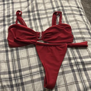 Röd bikini med spänne - Snygg röd bikini med ett dekorativt spänne framtill. Överdelen har breda axelband och en stilren design. Underdelen är enkel och matchar toppen perfekt. Perfekt för en dag på stranden eller vid poolen.