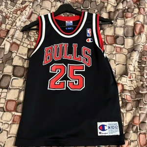 Chicago Bulls basketlinne Kerr #25 - Snyggt svart basketlinne från Champion med Chicago Bulls tryck och nummer 25, Kerr på ryggen. Linnet har röda och vita detaljer runt hals och ärmhål, samt stora tryck fram och bak. Perfekt för basketfans som vill sticka ut.