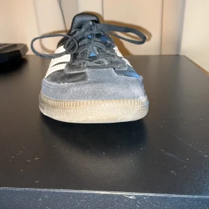 Adidas Samba svarta sneakers - Klassiska Adidas Samba sneakers i svart skinn med vita detaljer och de ikoniska tre ränderna på sidorna. Skon har mockadetaljer framtill, snörning och platt sula. Perfekt för dig som gillar retrostil och streetwear.