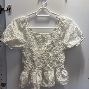 Vit blus med puffärm och volang - Supersöt vit blus med puffiga korta ärmar och smockad struktur. Toppen har fyrkantig ringning fram och v-ringning bak samt volangkant nertill. Perfekt för dig som gillar romantisk och trendig stil.