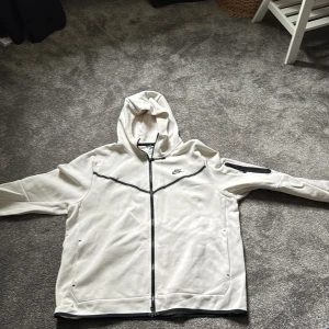 Vit Nike tech fleece  - Snygg vit Nike tech från Nike i storlek XL. Jackan har hel dragkedja, huva och svarta detaljer längs dragkedjan och bröstet. Praktiska fickor på sidorna och en extra ficka med dragkedja på ärmen. Perfekt för en sportig och avslappnad stil. Varm och skön säljer för jag ej använder den mer