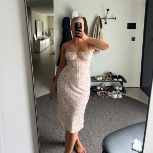 Blommig Adoore klänning  - Supersöt blommig klänning i ljusa rödaktiga färger, smala axelband med snörning och dessutom snörning framtill. Klänningen är midilång och har en figurnära passform med en dragkedja i ryggen. 