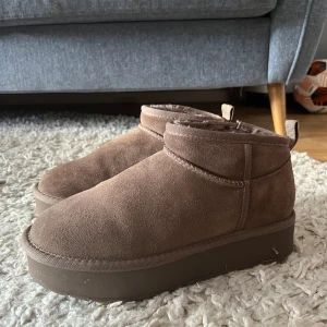 Bruna mockaboots från Pace, storlek 38 - Mysiga bruna boots i storlek 38 från Pace i mocka med fluffigt foder och platt, rejäl sula. Fodret i skorna är i ull, så de är perfekta till höst och vinter eftersom de håller värmen väldigt bra. Låg modell med dragflik bak för enkel påtagning. Lite slitna där bak men inget som förstör användningen av skorna. Använda några fåtals gånger. Nypris 749kr.