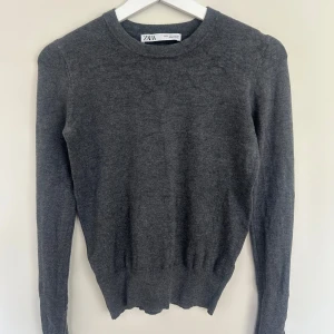 Mörkgrå stickad tröja från Zara - Mörkgrå stickad tröja från Zara med rund halsringning och långa ärmar. Tröjan har en enkel och clean look med ribbade muddar och små guldfärgade knappar vid ärmslutet.